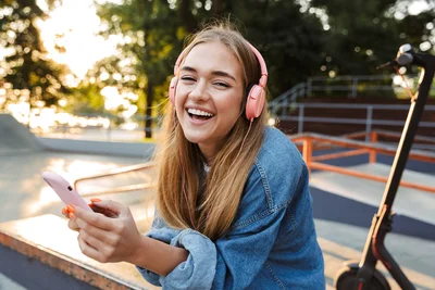 Eine junge Frau sitzt im Freien und schaut lächelnd auf ihr Smartphone; neben ihr steht ein Skateboard.